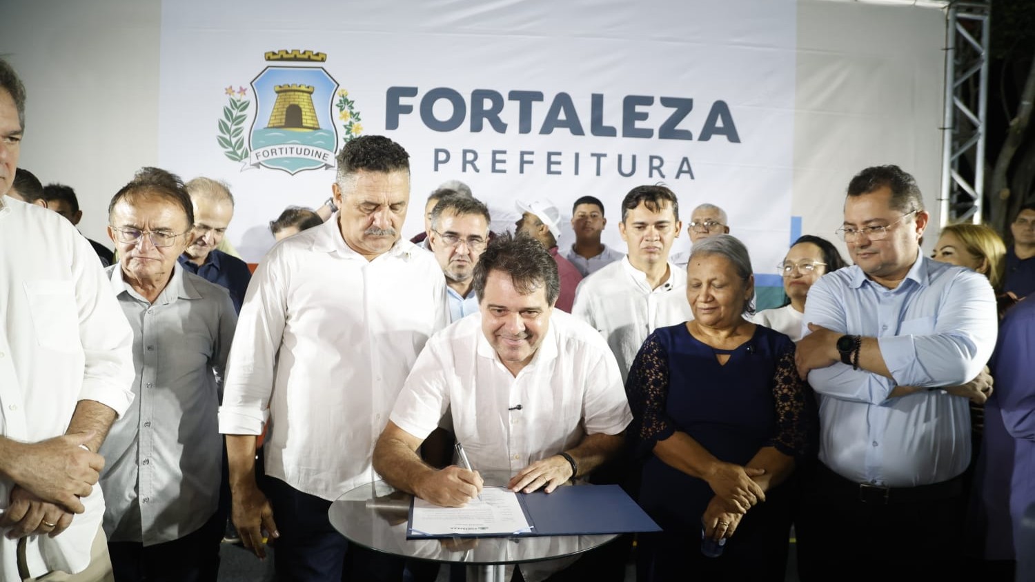 prefeito Evandro assinando um documento num palco, cercado por um grupo de pessoas